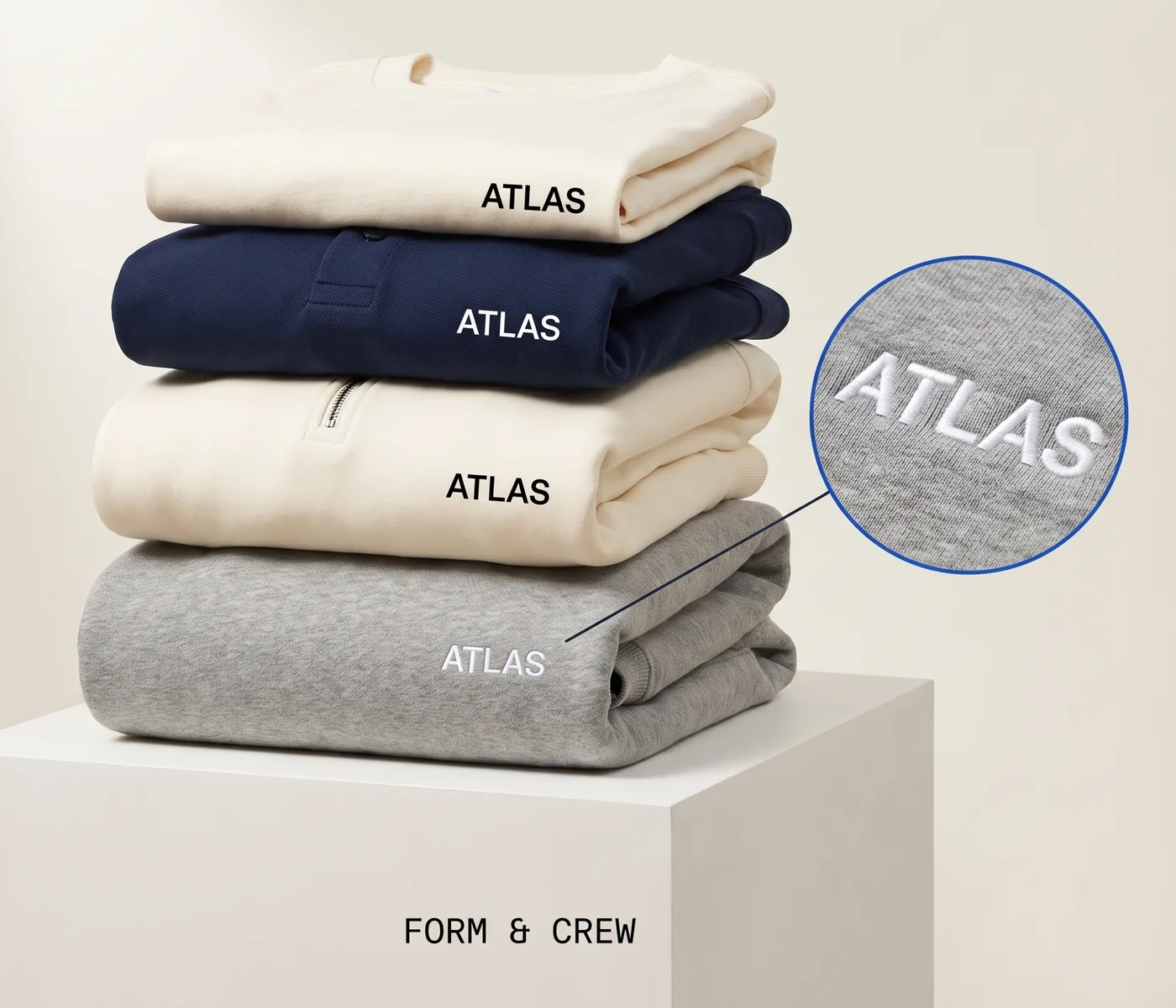 Folded stack of ATLAS garments — tee, navy polo, quarter-zip and grey crew — with embroidery detail